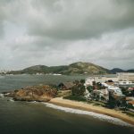 Vista aérea de uma área urbana litorânea do Espírito Santo, cercada por morros verdes sob céu nublado.