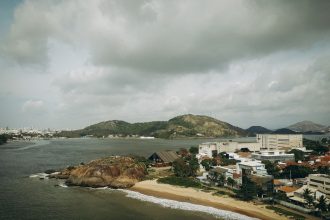Vista aérea de uma área urbana litorânea do Espírito Santo, cercada por morros verdes sob céu nublado.