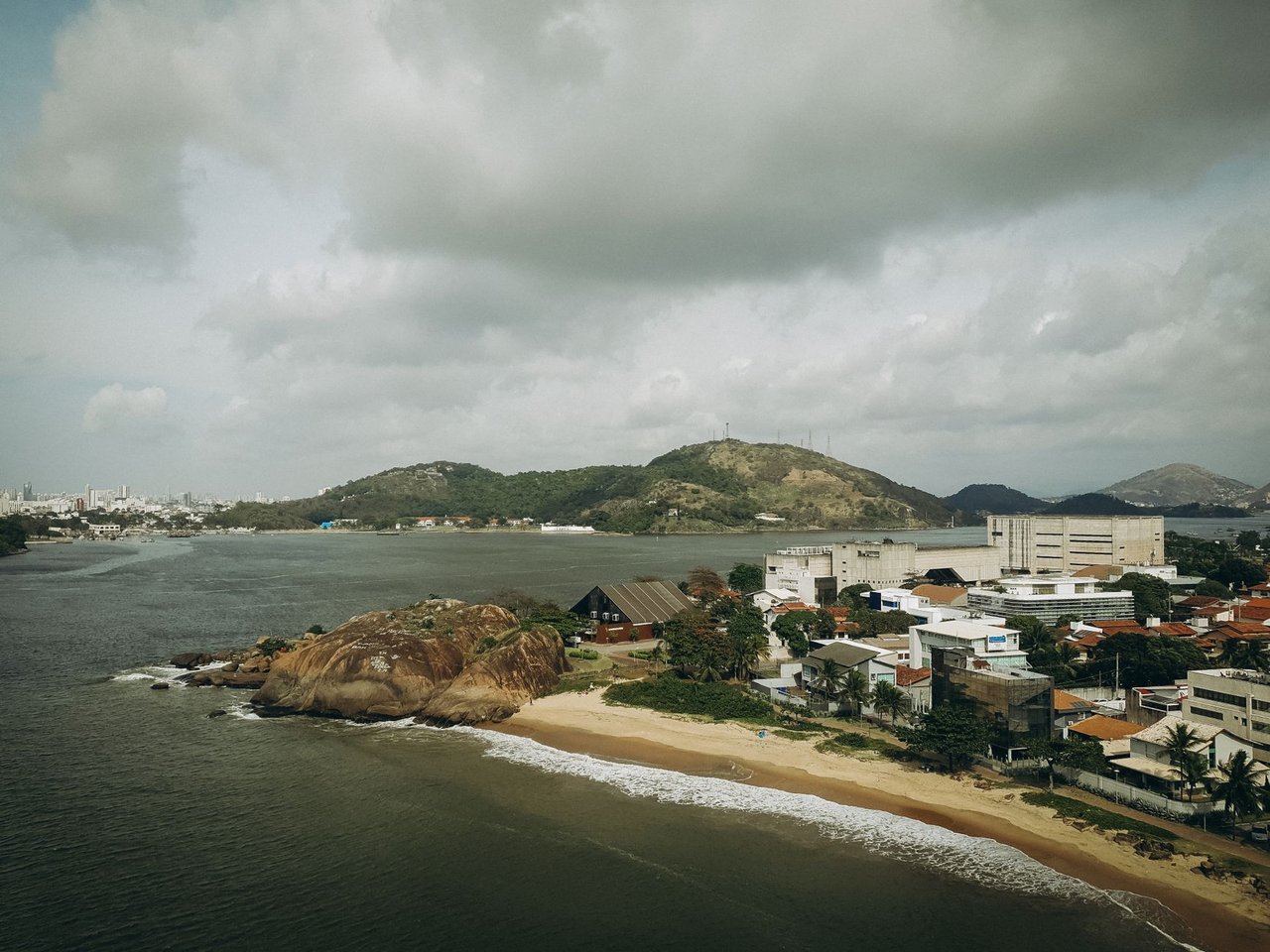 Vista aérea de uma área urbana litorânea do Espírito Santo, cercada por morros verdes sob céu nublado.