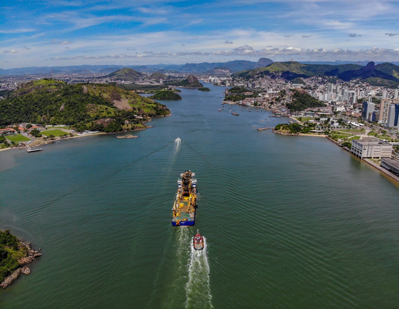 Vista aérea de Vitória, no Espírito Santo, com a orla portuária e um grande navio navegando pelo canal.