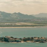 Vista aérea do litoral de Vitória, com áreas urbanas, mar e montanhas ao fundo.