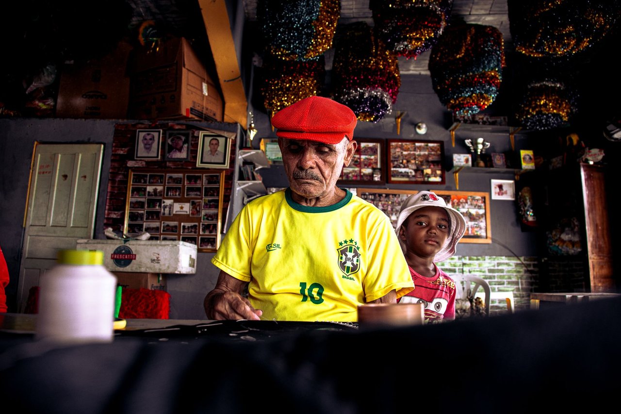 Homem idoso e menino criam uma peça artística juntos em uma oficina colorida no Brasil.