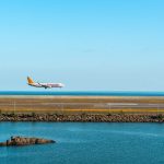 Avião comercial se aproxima de uma pista de pouso à beira-mar sob céu azul.